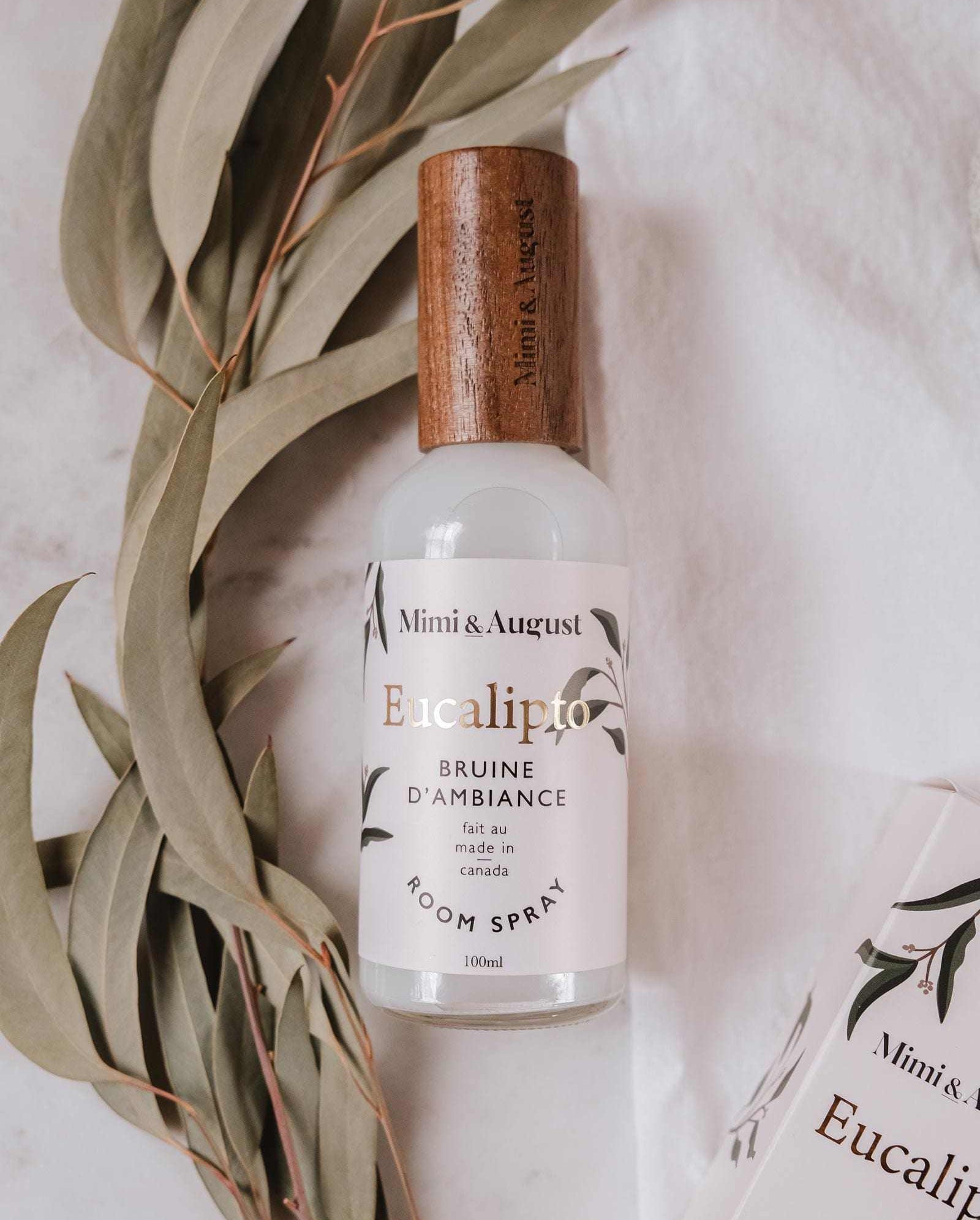 A bottle of Mimi & August Eucalypto Room Spray with a wooden cap sits beside fresh eucalyptus leaves on a light surface, emphasizing its natural fragrance.