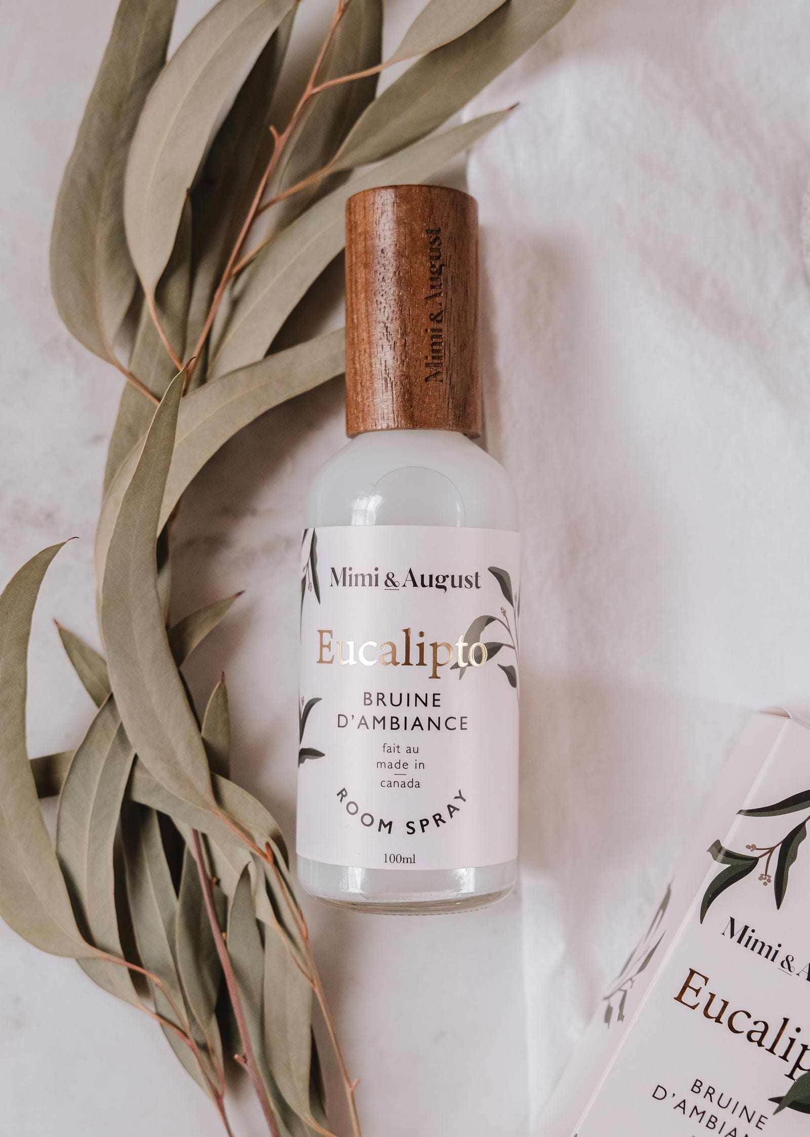 Une bouteille de Bruine d'Ambiance Eucalyptus Mimi & August, munie d’un capuchon en bois, est posée à côté de feuilles d’eucalyptus fraîches sur une surface claire, mettant en valeur son parfum naturellement frais.