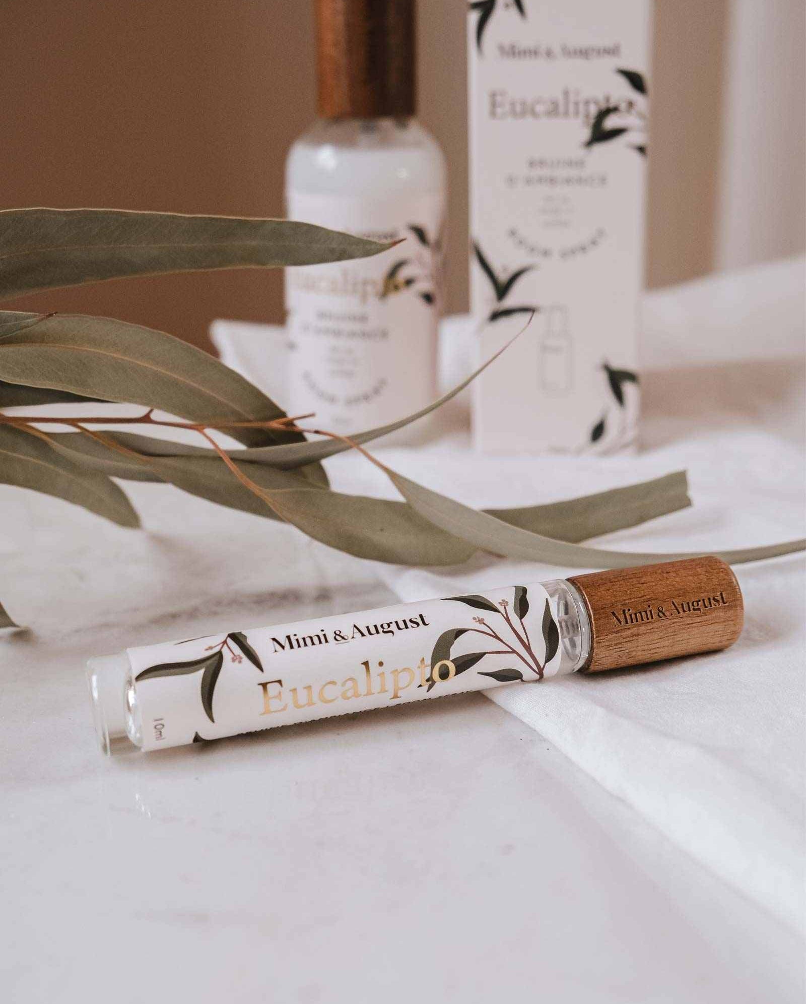 A bottle of Mimi & August Eucalypto Room Spray rests on a white surface, surrounded by fresh eucalyptus leaves and packaging, capturing the refreshing essence of its eucalyptus scent.