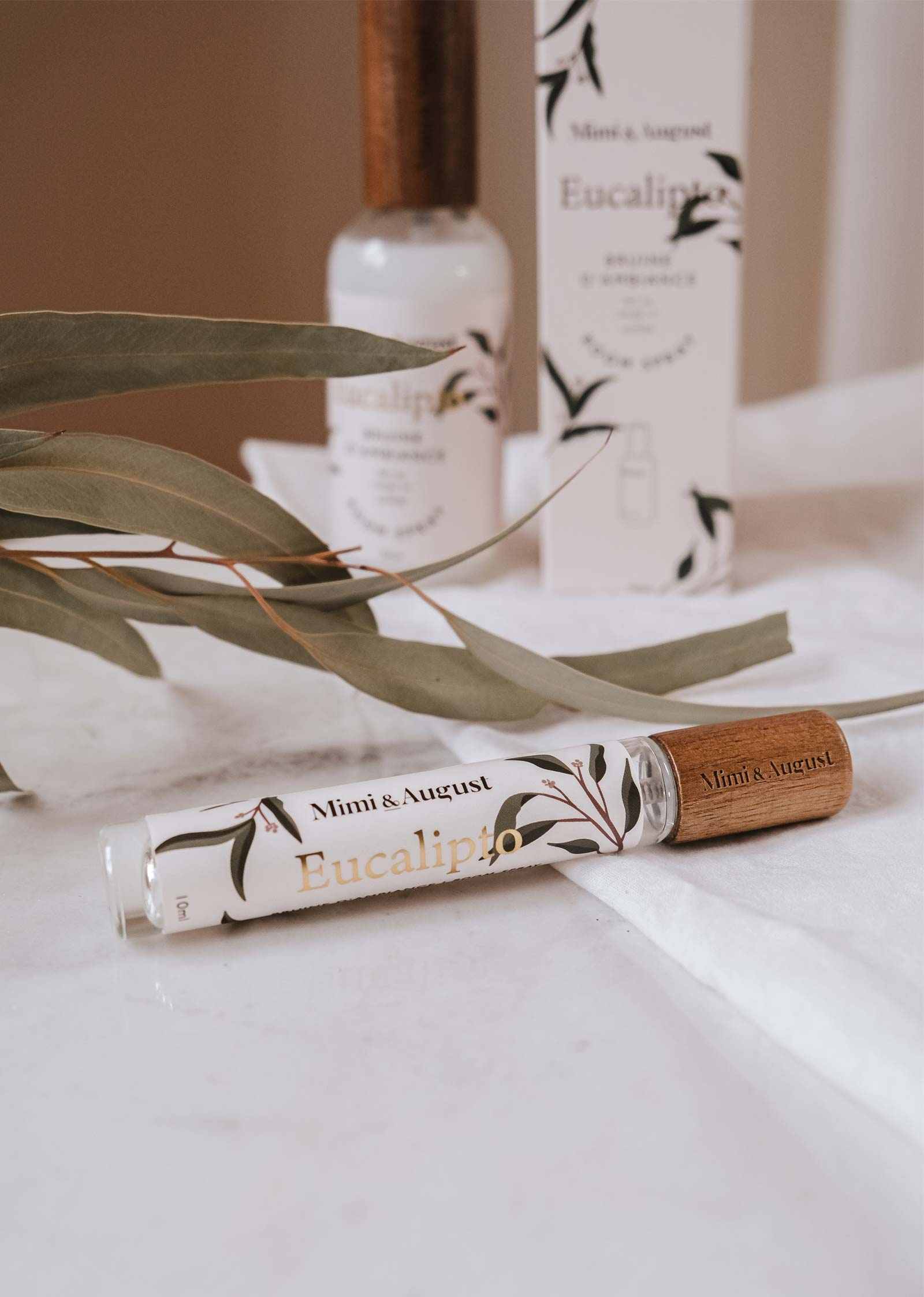Une bouteille de Bruine d'Ambiance Eucalyptus Mimi & August repose sur une surface blanche, entourée de feuilles d’eucalyptus fraîches et de son emballage, capturant toute l’essence rafraîchissante et vivifiante de l’eucalyptus.