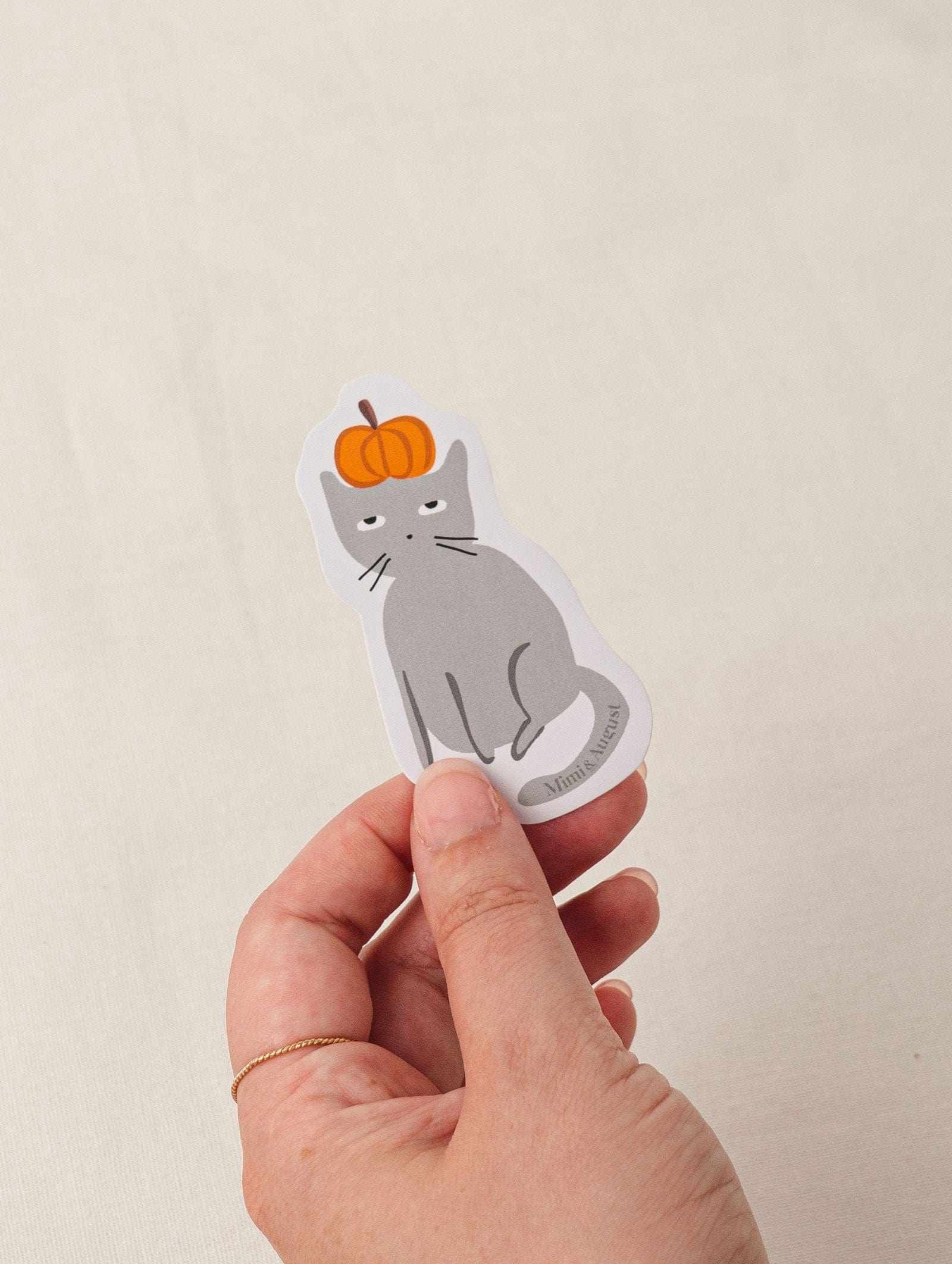 Hand holding Sacha the Pumpkin Lover vinyl sticker featuring gray cat with pumpkin on head
