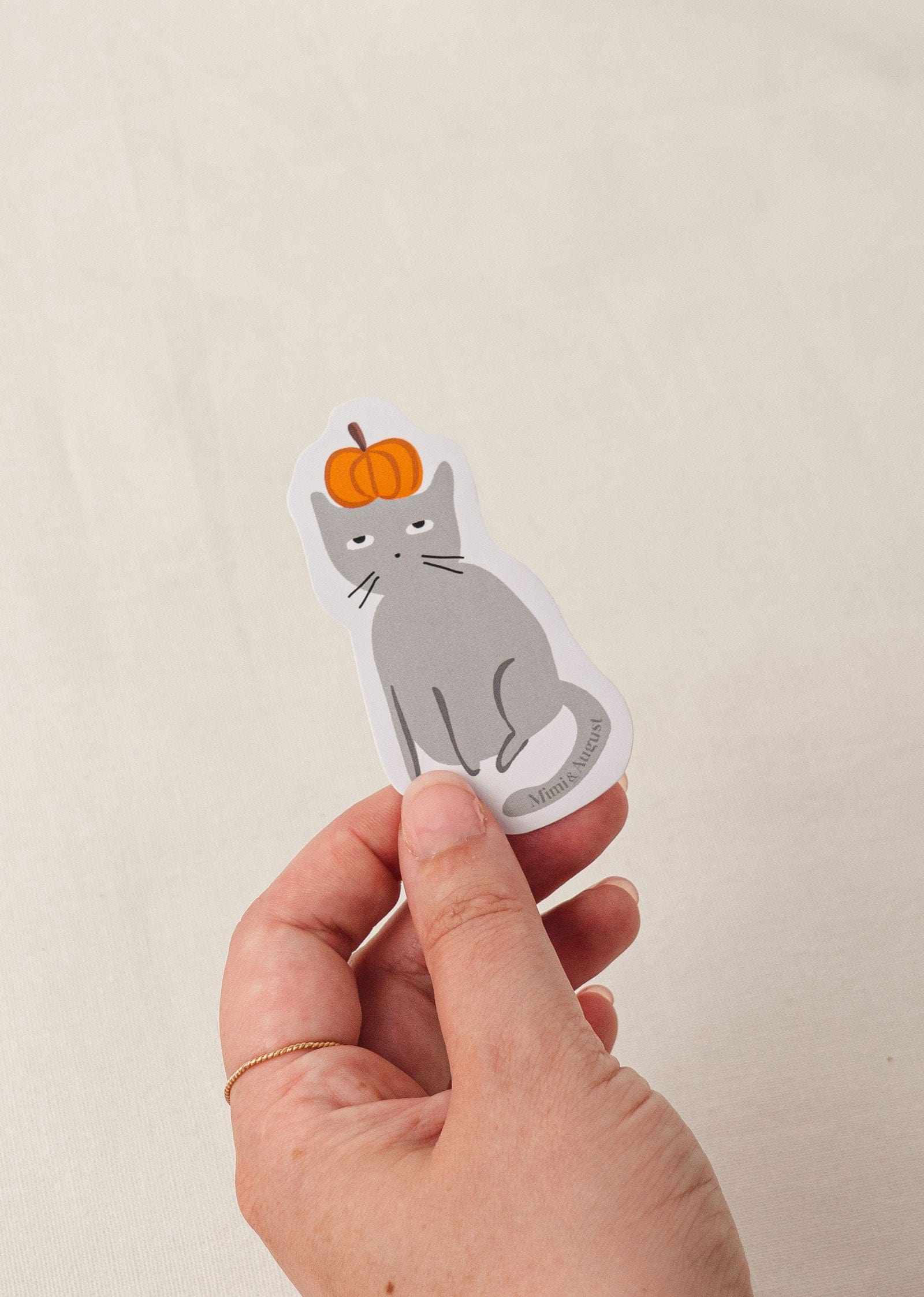Hand holding Sacha the Pumpkin Lover vinyl sticker featuring gray cat with pumpkin on head