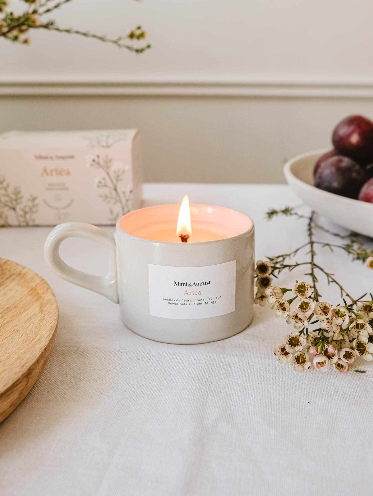 The Artea – Reusable Soy Candle by Mimi & August glows in a mug on a table, surrounded by a box, a bowl of plums, a wooden plate, and sprigs of small white flowers.