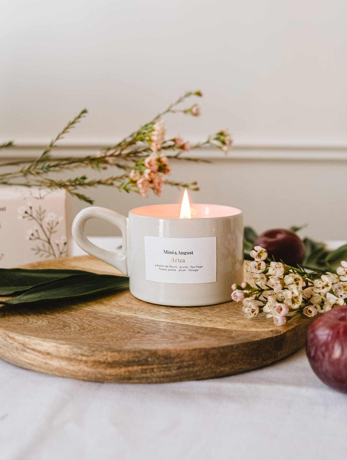 The Artea – Reusable Soy Candle by Mimi & August glows in a ceramic holder atop a wooden tray, surrounded by delicate flowers and leaves, with a plum and a floral box in the background.