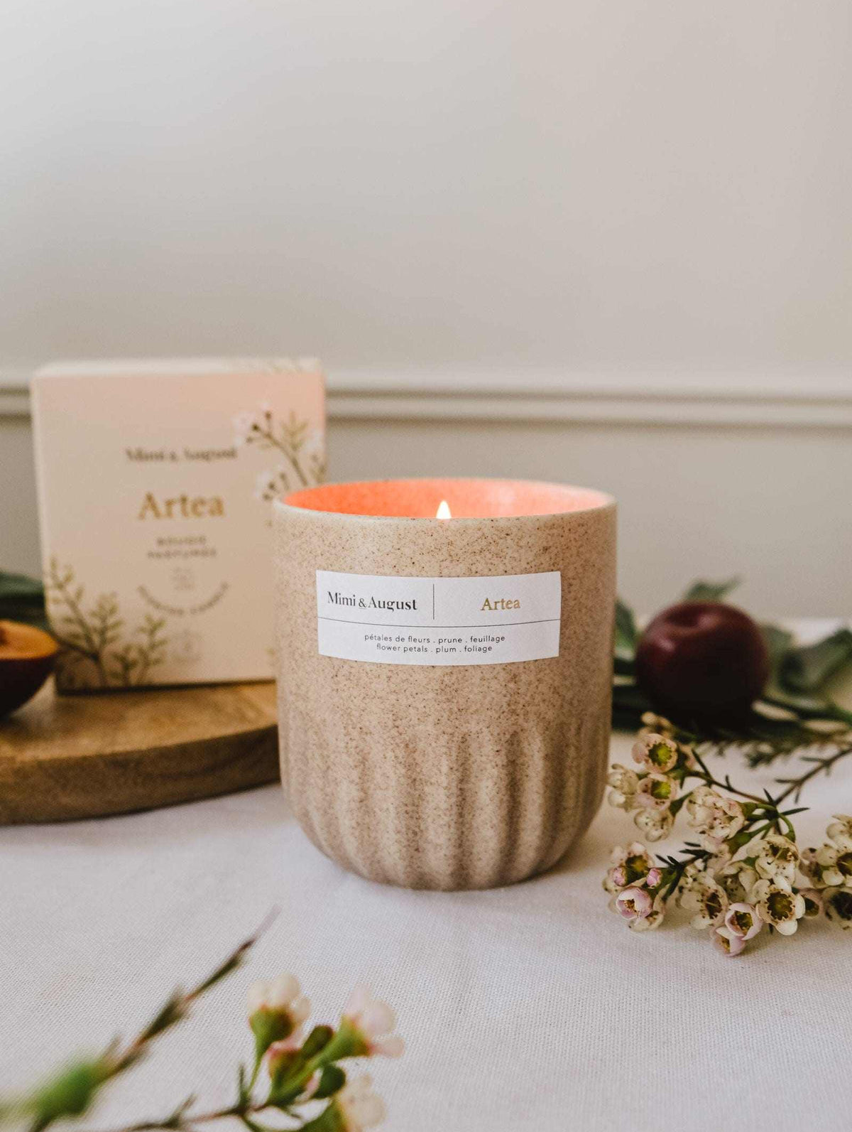 A beige floral Mimi & August Artea – Reusable Soy Candle is lit on a table with small flowers, greenery, and its box in the background, adding charm and sustainability to any space.