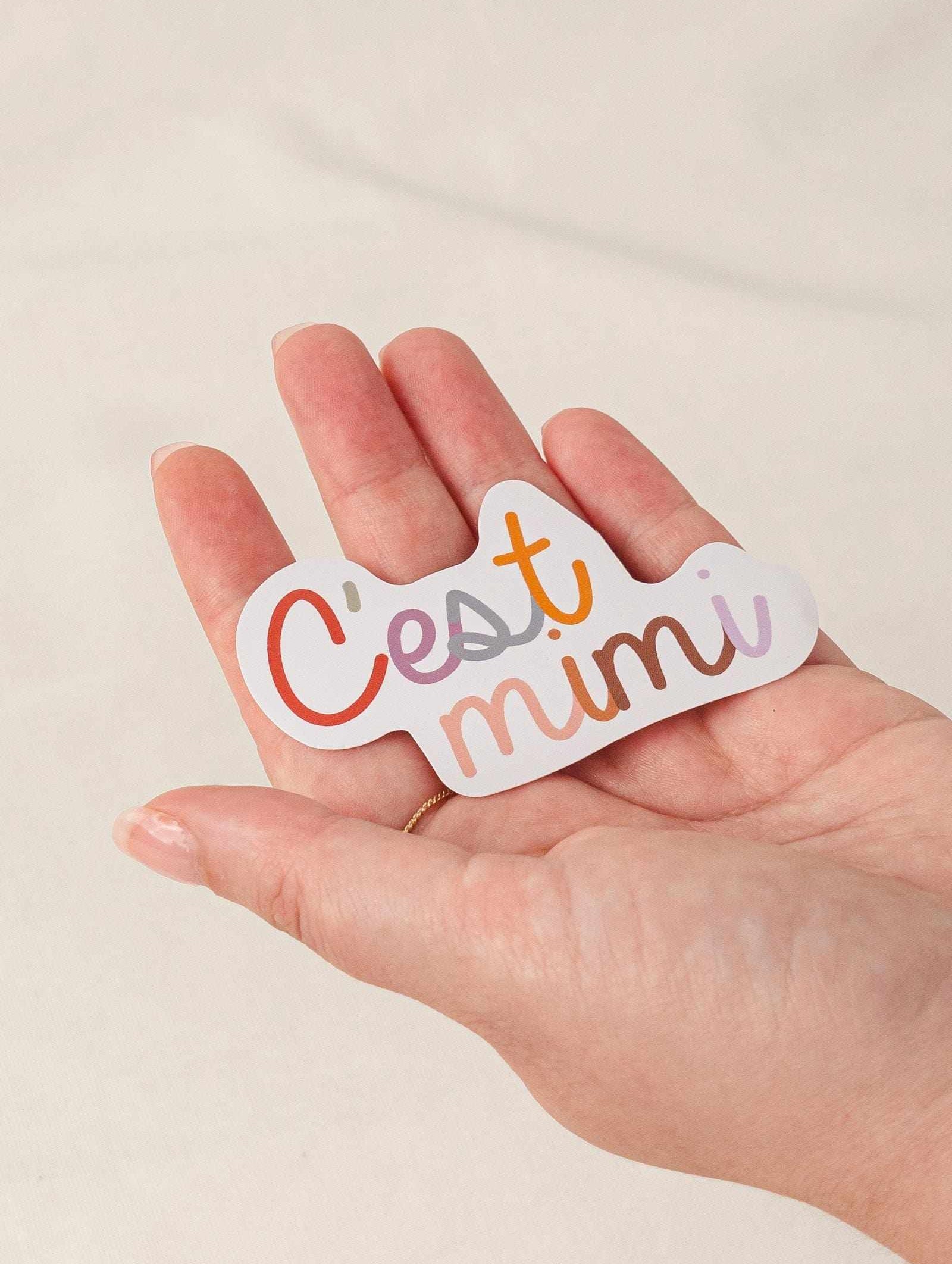 Hand holding a water-resistant vinyl sticker with "C'est mimi" text and white border