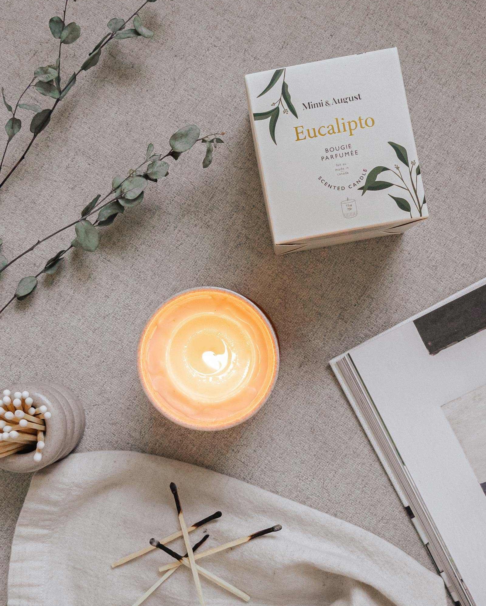 Eucalyptus scented reusable candle with minty and woody fresh aroma, shown lit next to its box and matches on a cozy fabric surface
