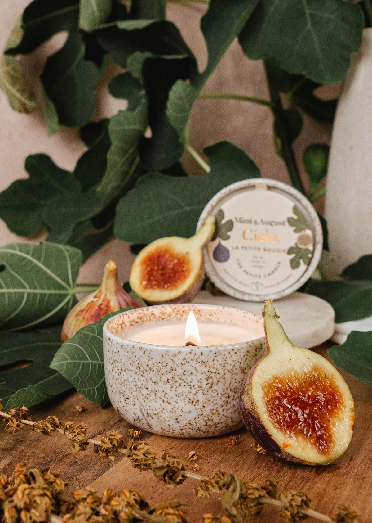 Burning fig candle in reusable ceramic cup surrounded by fresh figs and green leaves
