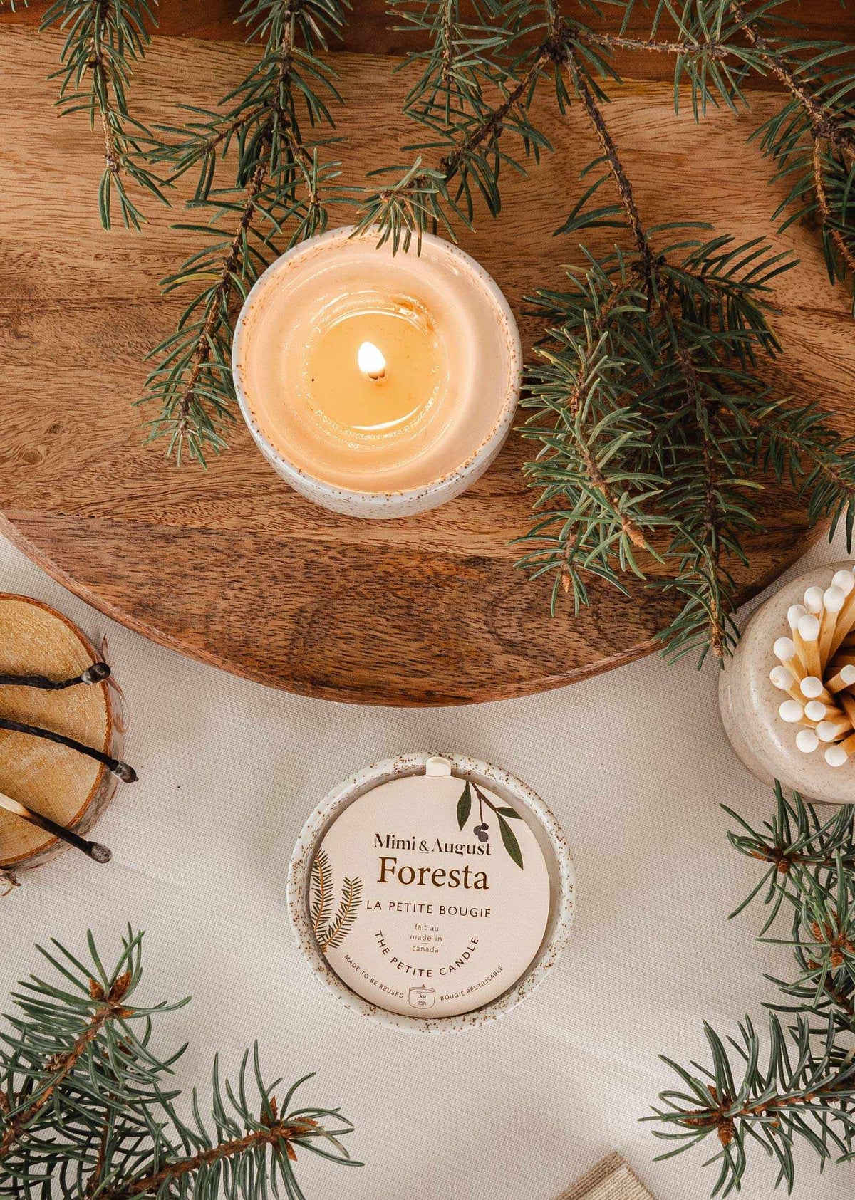 Lit fir tree candle in reusable ceramic cup surrounded by fir branches and candle lid on wooden surface