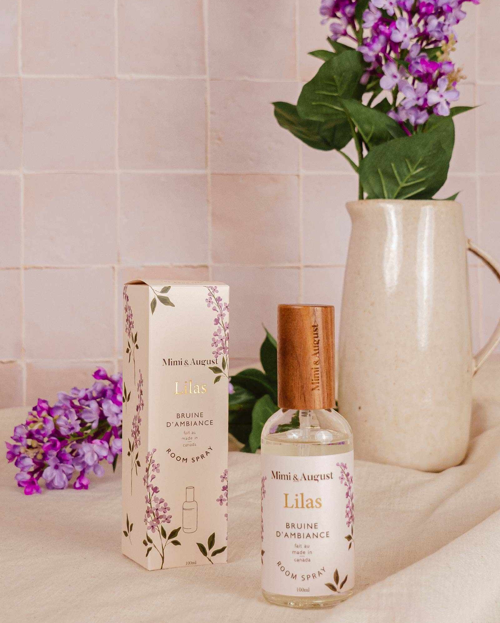 A bottle of Mimi & August's Room Spray - Lilas and its box sit on a beige surface, with a ceramic pitcher and purple flowers in the background.