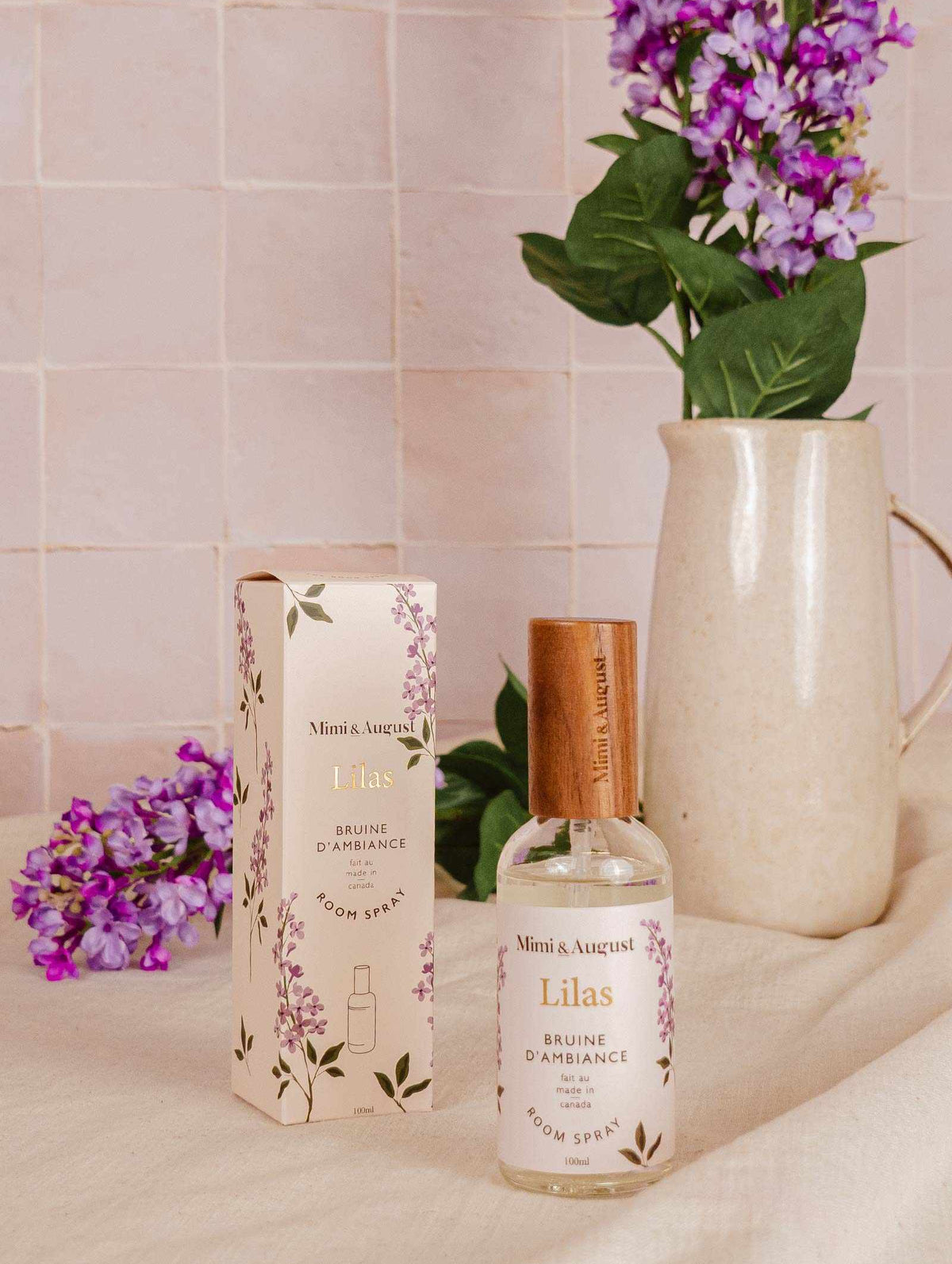 A bottle of Mimi & August's Room Spray - Lilas and its box sit on a beige surface, with a ceramic pitcher and purple flowers in the background.