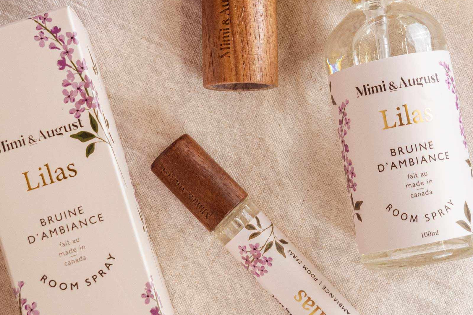 Room spray - Lilas by Mimi & August and packaging on a fabric surface with purple flowers. Includes a boxed spray, an uncapped spray bottle, a woody cap, and a small spray vial. Labeled "Mimi & August Lilas," this Room spray - Lilas features premium fragrance oil for an exquisite scent experience.