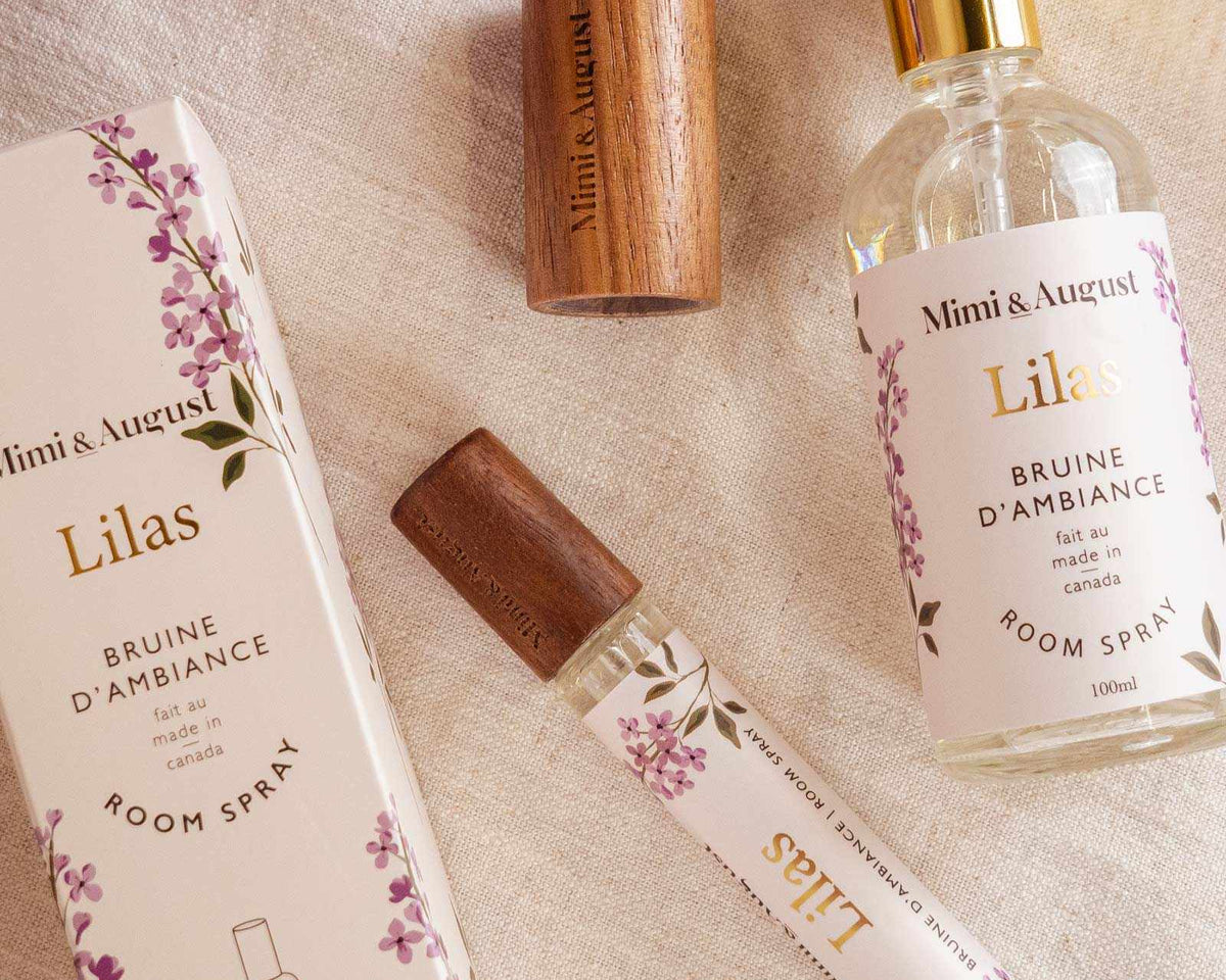 Room spray - Lilas by Mimi & August and packaging on a fabric surface with purple flowers. Includes a boxed spray, an uncapped spray bottle, a woody cap, and a small spray vial. Labeled "Mimi & August Lilas," this Room spray - Lilas features premium fragrance oil for an exquisite scent experience.