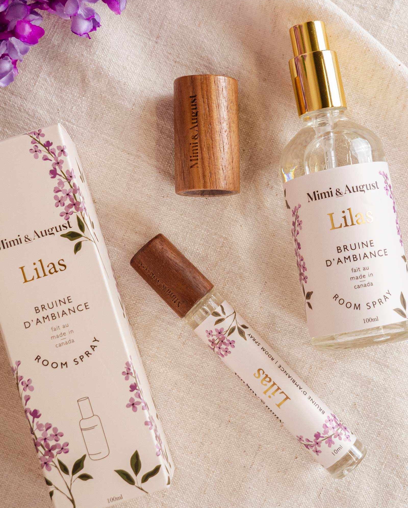 Room spray - Lilas by Mimi & August and packaging on a fabric surface with purple flowers. Includes a boxed spray, an uncapped spray bottle, a woody cap, and a small spray vial. Labeled "Mimi & August Lilas," this Room spray - Lilas features premium fragrance oil for an exquisite scent experience.