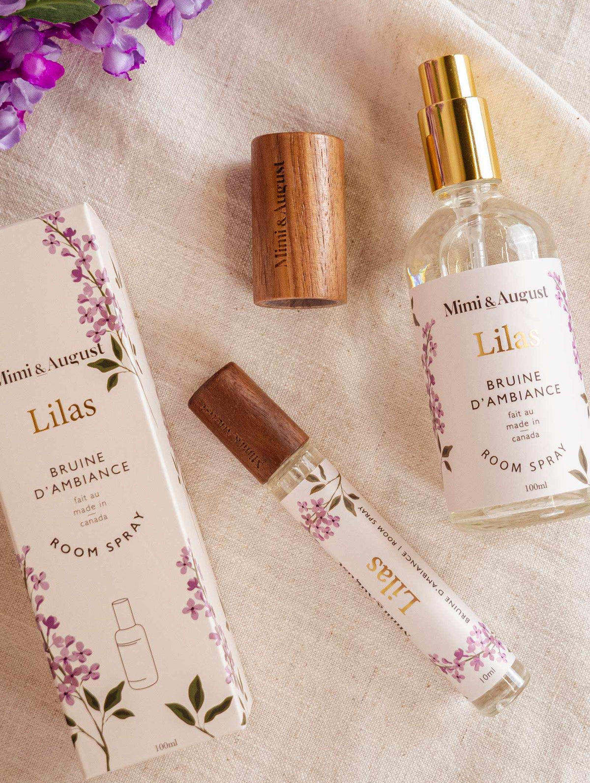 Room spray - Lilas by Mimi & August and packaging on a fabric surface with purple flowers. Includes a boxed spray, an uncapped spray bottle, a woody cap, and a small spray vial. Labeled "Mimi & August Lilas," this Room spray - Lilas features premium fragrance oil for an exquisite scent experience.