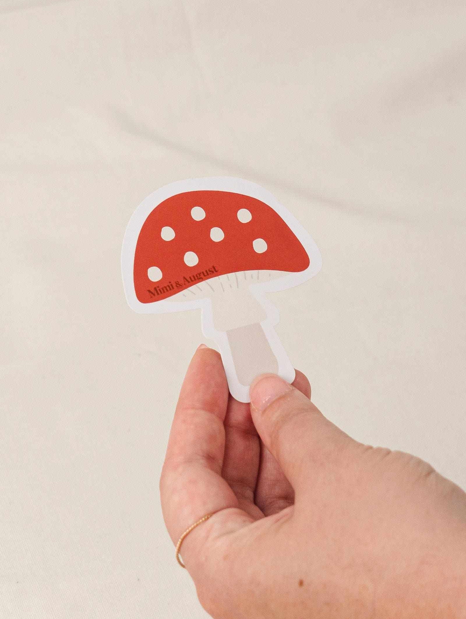 Hand holding a red and white polka dot mushroom vinyl sticker with white border on a beige background