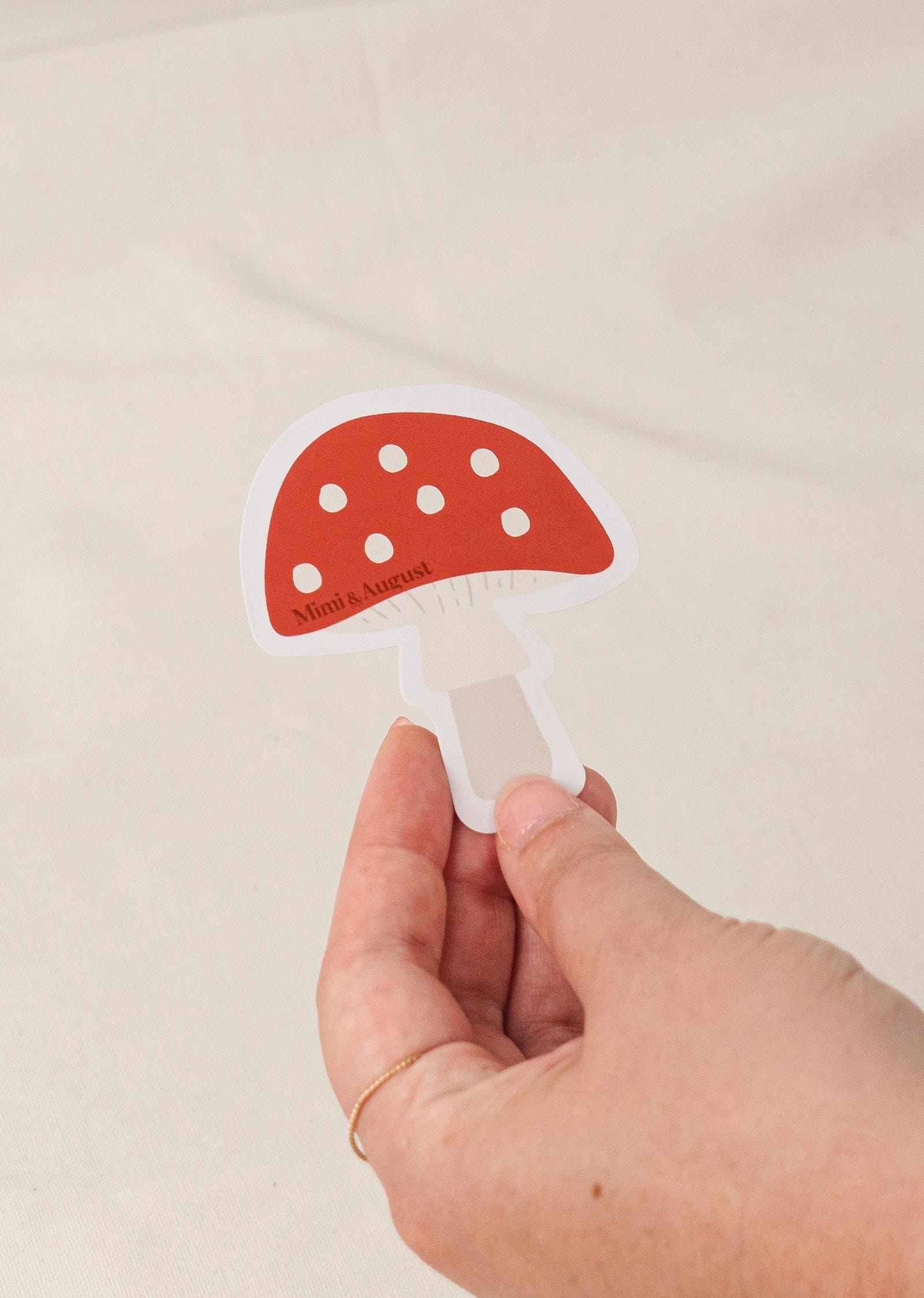 Hand holding a red and white polka dot mushroom vinyl sticker with white border on a beige background