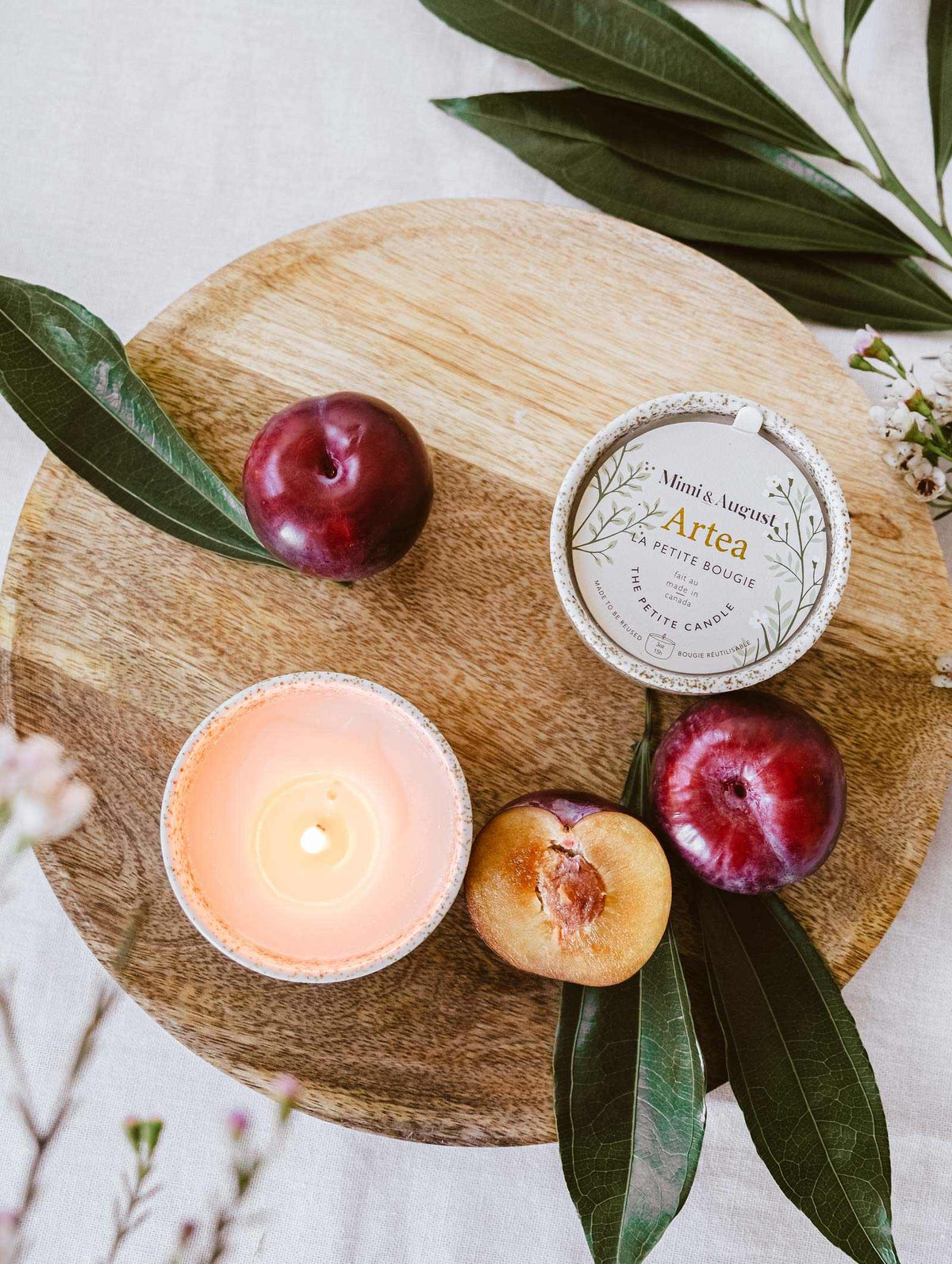 A lit Mimi & August Artea – Reusable Soy Candle in a ceramic container, plums, a halved plum, and the candle’s tin are arranged on a round wooden board with green leaves.