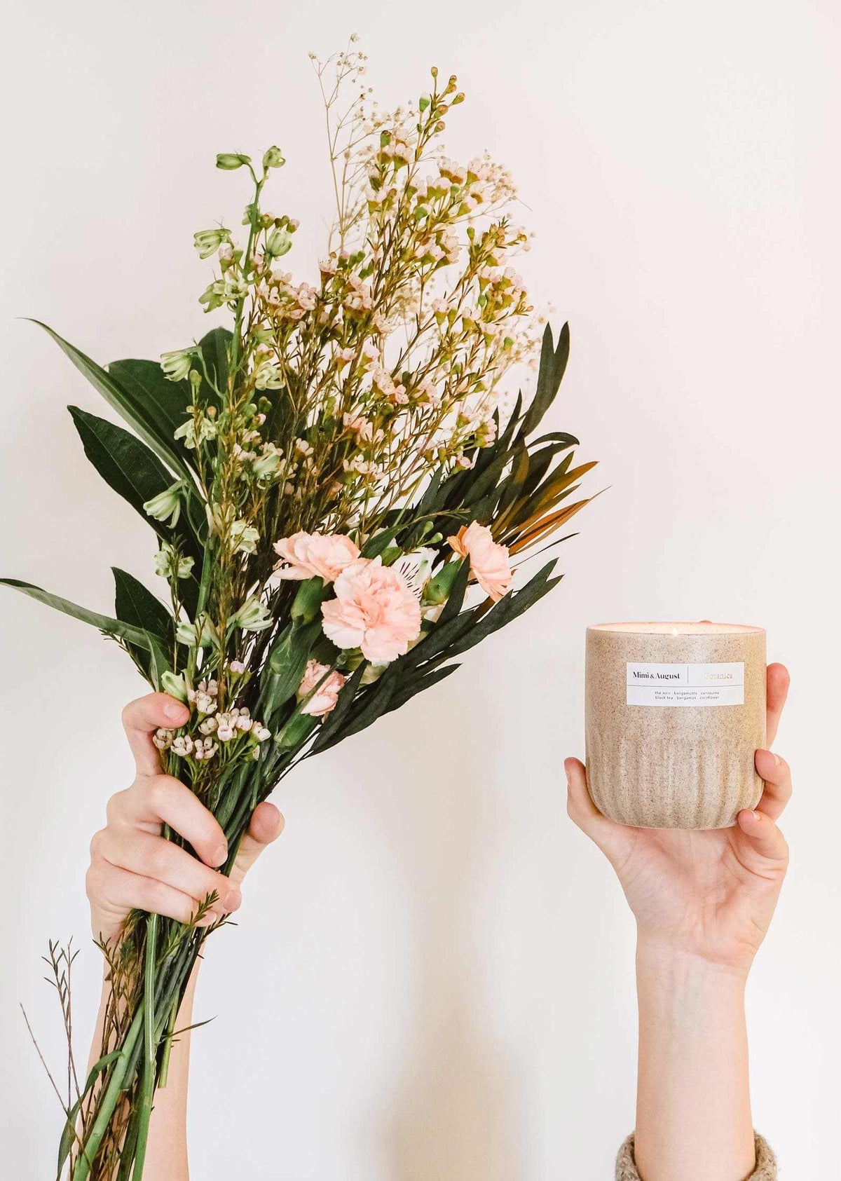 Des mains tenant un bouquet de fleurs roses et une bougie de soya réutilisable Botanica de Mimi & August sur un fond neutre.