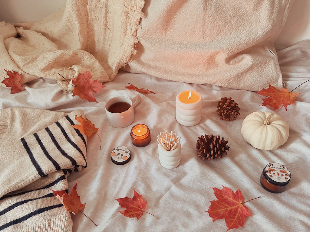Mimi & August exlusive fall candles sitting on a warm cozy fall bed beside a sweater, a blanket, a pillow, a warm cup of tea, fall leaves and a white pumkin