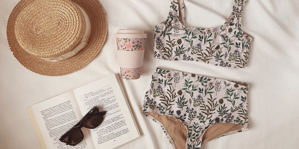 Herboria Maillot de bain et tasse de café de Mimi & August allongées sur un lit avec des accessoires d'été