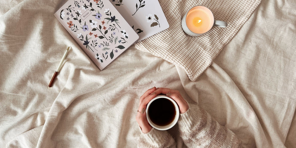 Des mains tenant une tasse de thé sur un lit, à côté de cahiers et d'une bougie Botanica de Mimi & August.