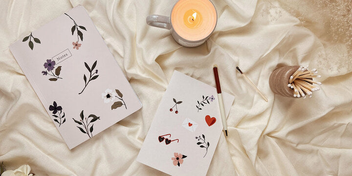 Carnet de notes à fleurs, carte de la Saint-Valentin et bougie parfumée à la tasse de Mimi & August posés sur une couverture confortable.