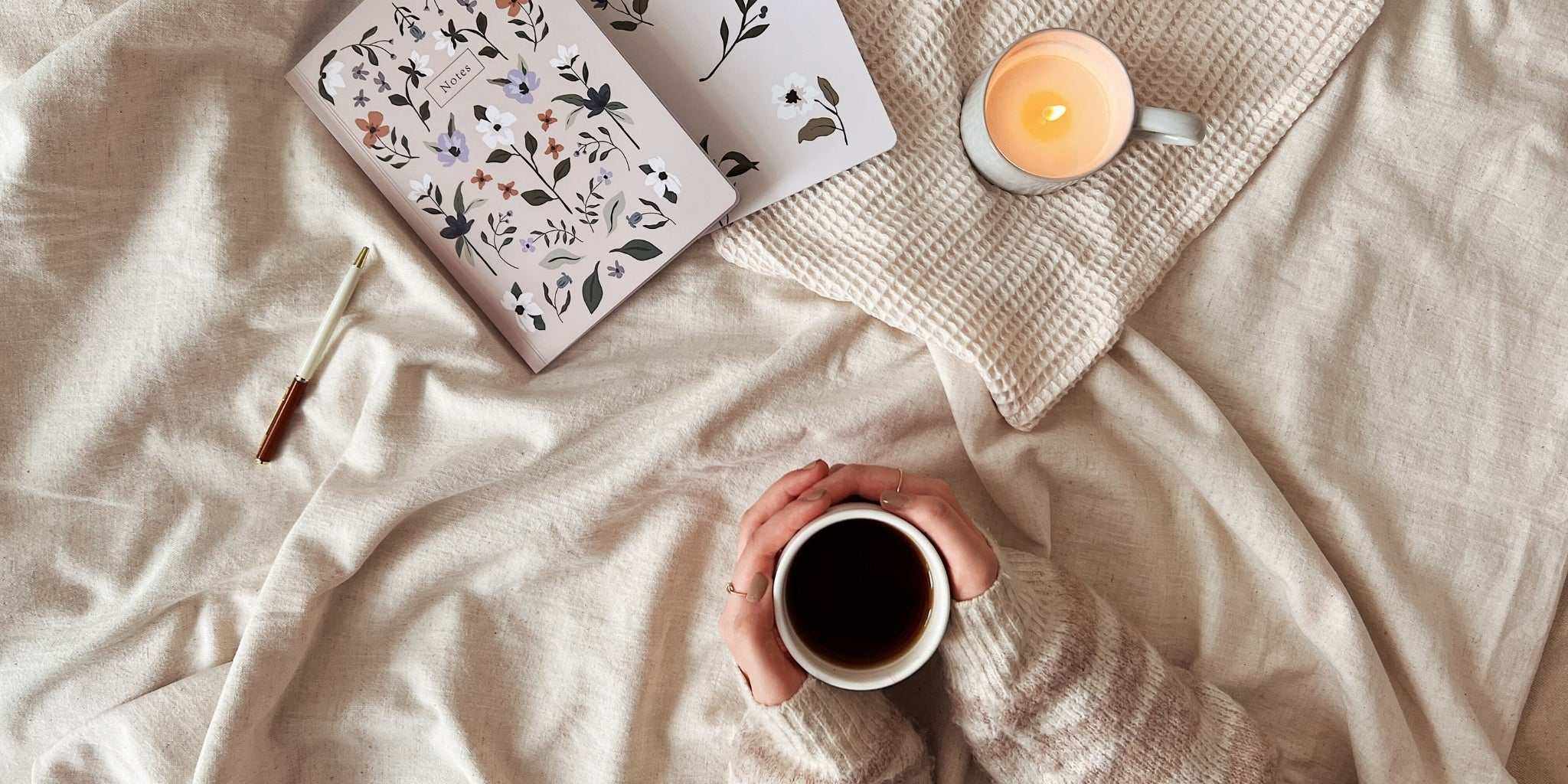 Hands holding a white ceramic mug filled with black coffee on a beige fabric surface with a lit candle and floral notebook for winter self-care tips