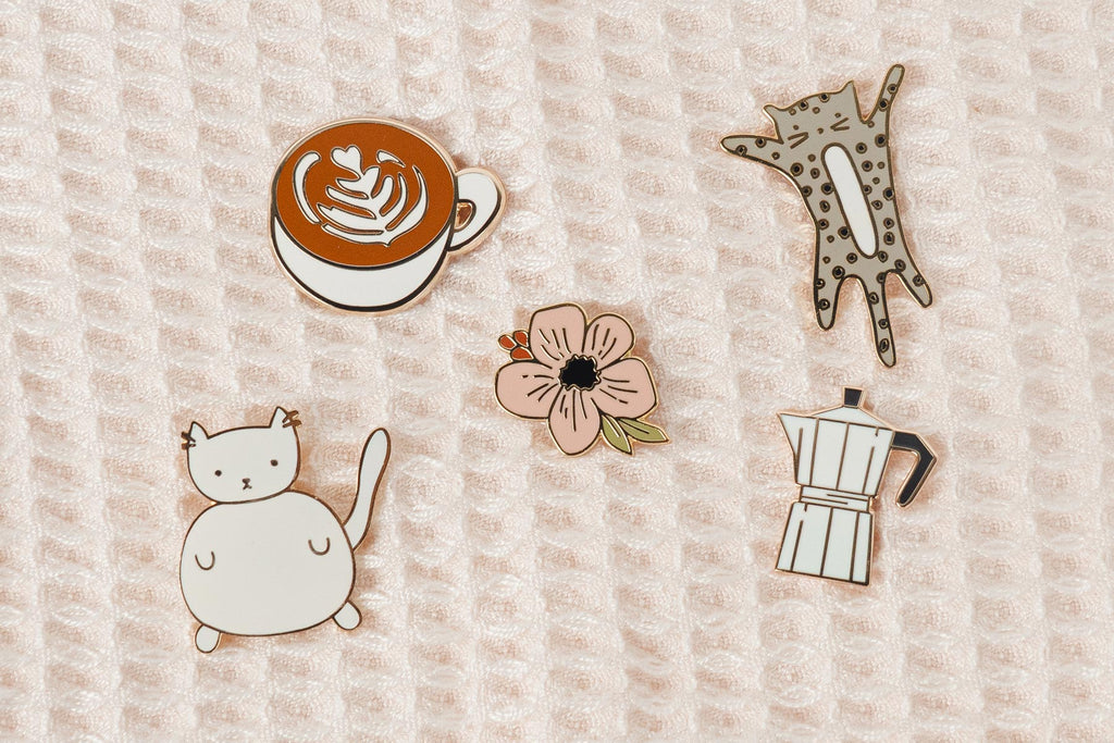 A set of enamel pins with a cat, a flower and a cup of coffee.