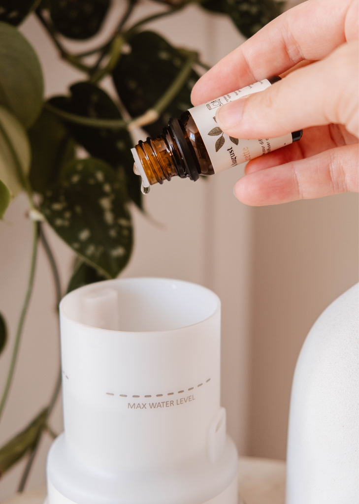 A hand adding Mimi & August Amalfi scented oil to a white diffuser, surrounded by tropical foliage.