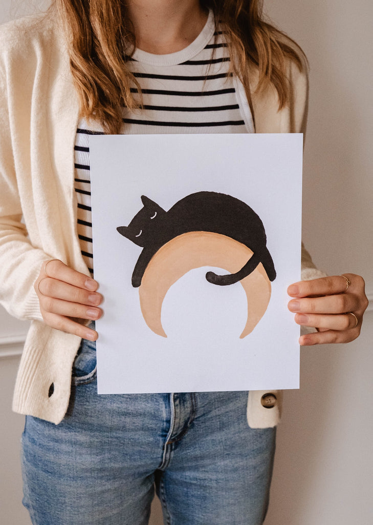 Une femme brandissant une illustration d'un chat noir sur un croissant de lune par Mimi & August.