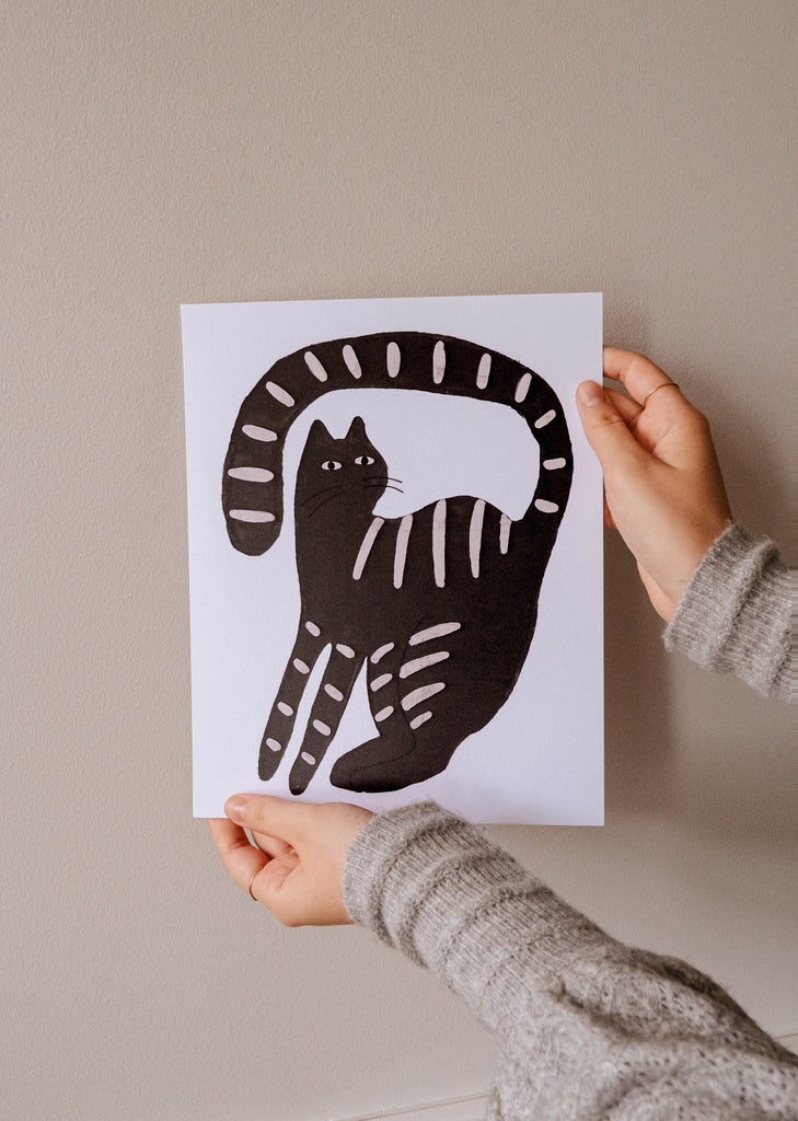 Une personne brandissant une illustration d'un chat noir calme de Mimi & August.
