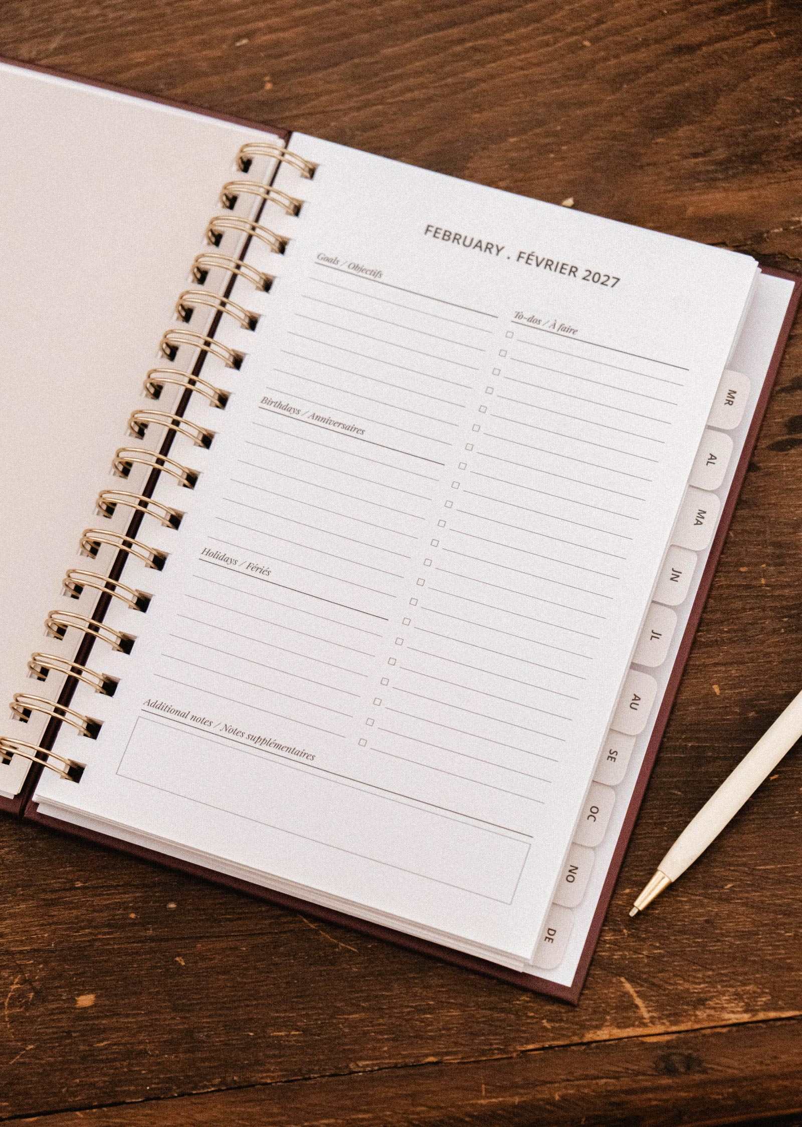 The Blooming Branch – 2027 Agenda by Mimi & August rests open on a wooden desk, displaying the February spread with dedicated spaces for goals, birthdays, holidays, and notes. A pen sits nearby, while delicate blooming branch illustrations enhance the planner’s charming and organized layout.