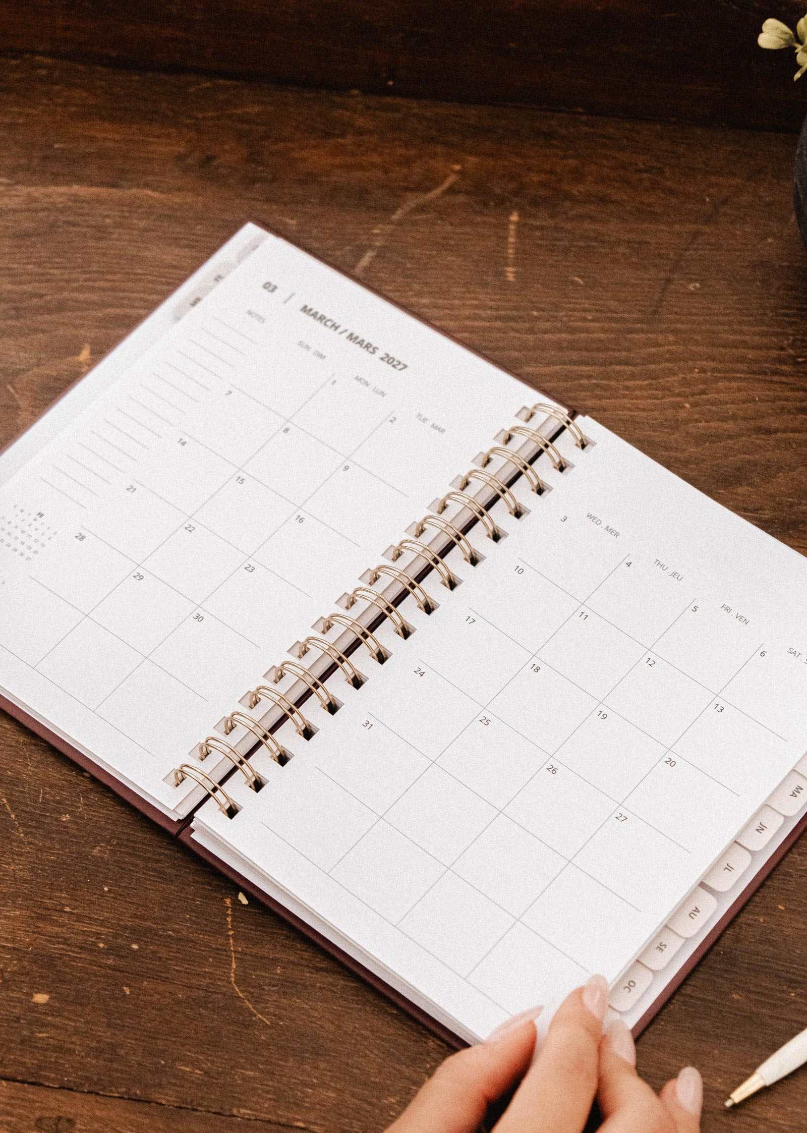 A Mimi & August Blooming Branch – 2027 Agenda, an open spiral-bound illustrated planner displaying a monthly calendar, rests on a wooden desk with a person's hand visible in the corner.