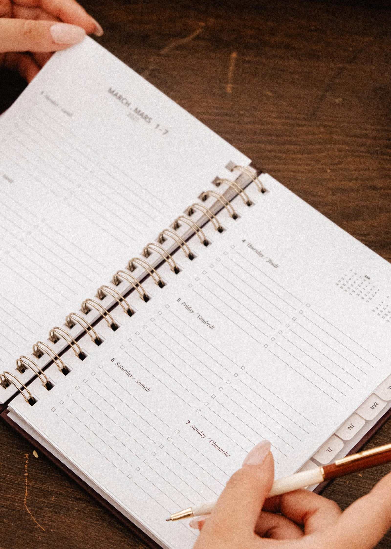 A person holding a pen opens the Mimi & August Wildflowers – 2027 Agenda, a spiral-bound illustrated planner, to a blank page labeled March 1-7 with sections for each day of the week.