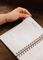 A hand with light pink nails holds open the Mimi & August Blooming Branch – 2027 Agenda, a spiral-bound illustrated planner showing a weekly schedule for March 1–7, on a wooden surface.