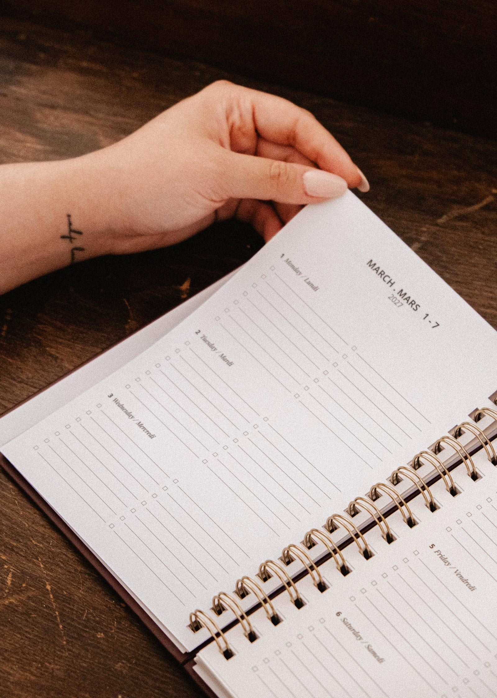 A hand with light pink nails holds open the Mimi & August Blooming Branch – 2027 Agenda, a spiral-bound illustrated planner showing a weekly schedule for March 1–7, on a wooden surface.