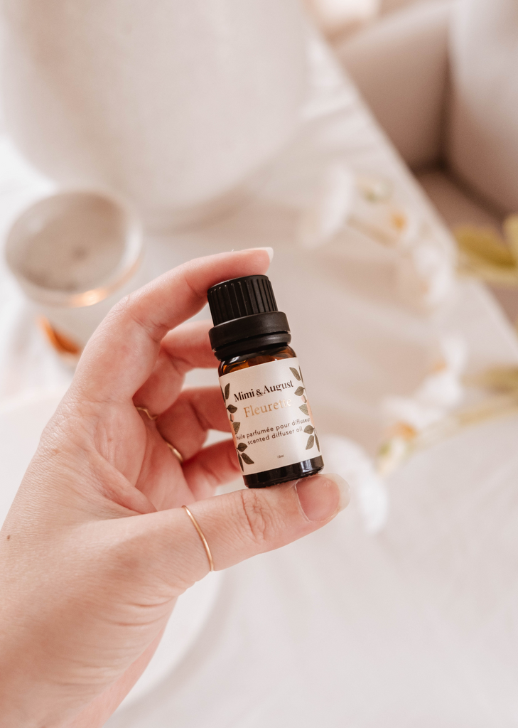 A hand holding a small bottle of essential oil labeled "Fleurette scented oil" by Mimi & August, with a blurred background featuring soft white tones and candles.