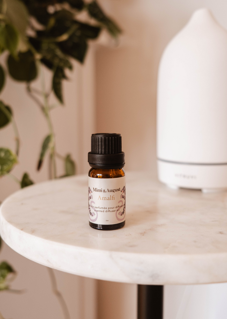 A small brown glass bottle of Amalfi scented oil for diffuser sits on a marble side table next to a white, modern diffuser, surrounded by tropical foliage.
