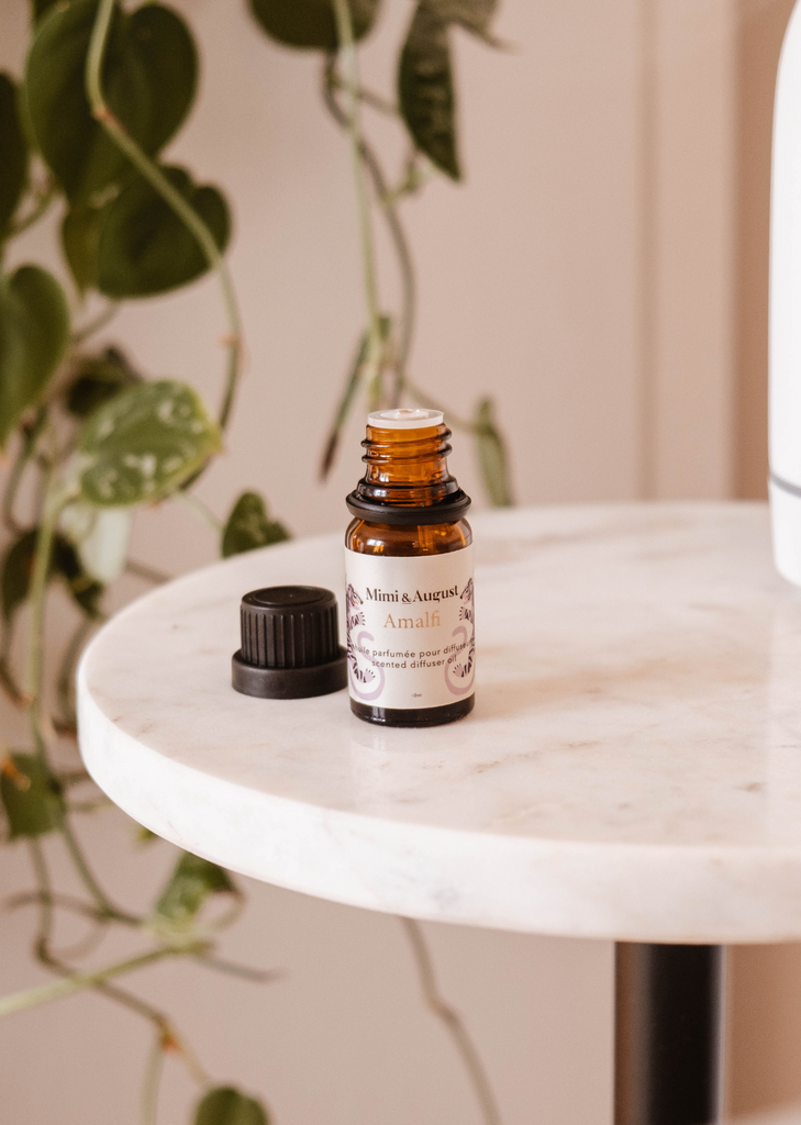 Small amber glass bottle of Amalfi scented oil for diffuser with a black cap beside it, resting on a marble table next to potted tropical foliage.