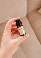 A hand holding a small bottle of Mimi & August's Confetti scented oil in front of a beige sofa.