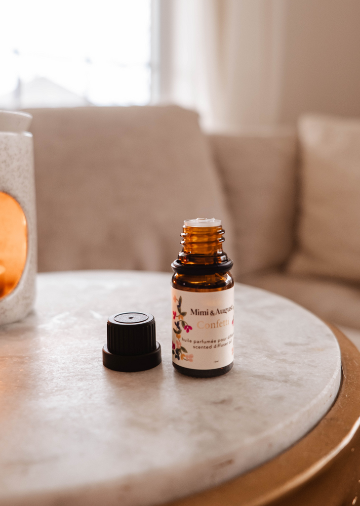 A small bottle of Mimi & August Mandarin Confetti scented oil on a marble tabletop with its cap removed, beside a lit candle, with a cozy sofa in the background.