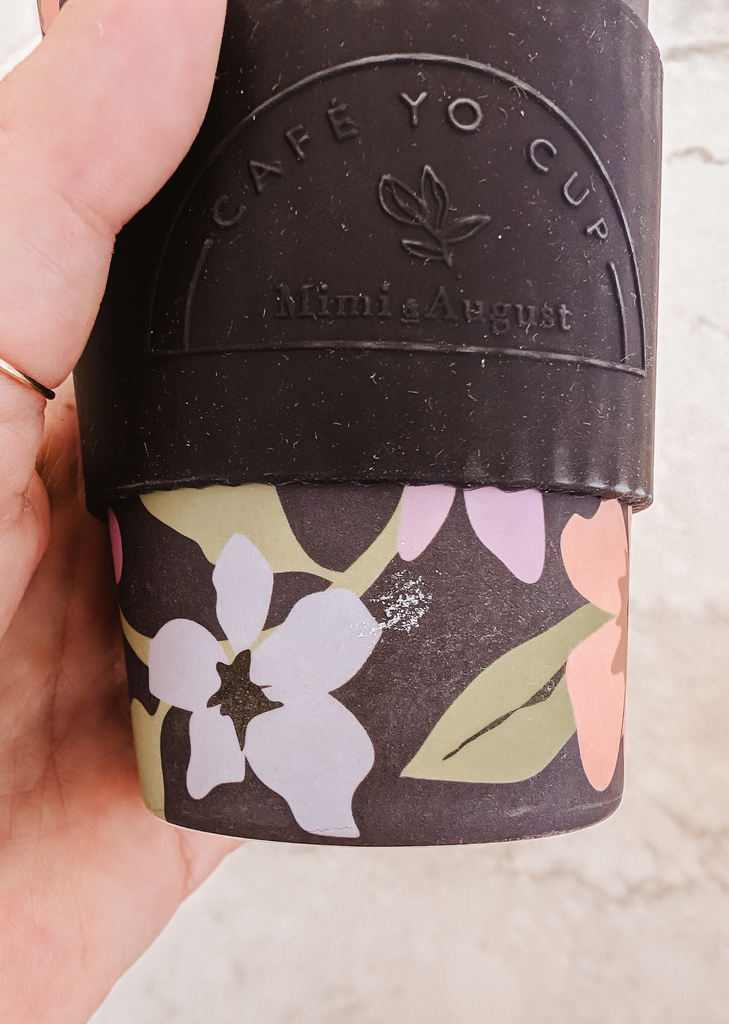 A person holding up a Defect Café Yo Cup by Mimi & August with flowers and a silicone lid on it.