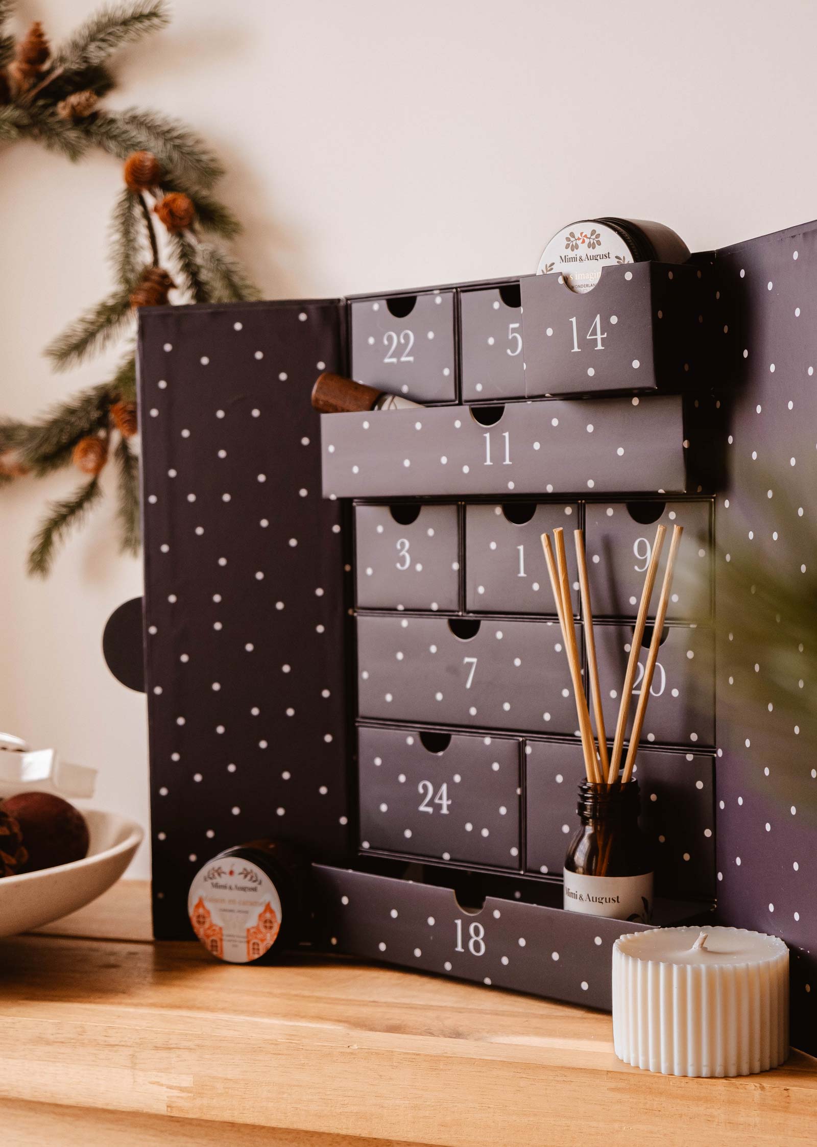 Le Calendrier de l'Avent - Les Douze Jours Enchantés de Mimi & August, avec son design à pois noirs et blancs et ses tiroirs ouverts, est élégamment présenté sur une surface en bois. Il est entouré de décorations de Noël et accompagné d'une bougie parfumée, dont la chaude fragrance renforce le charme festif.