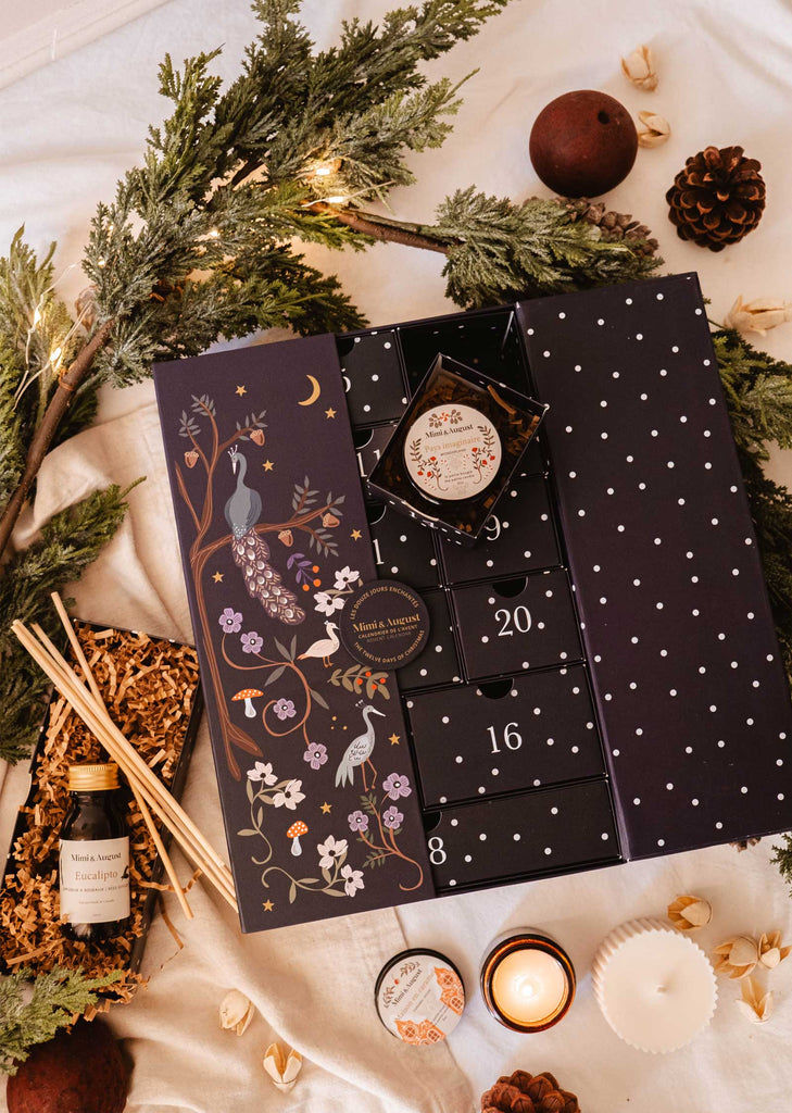 The Advent calendar from Mimi & August, called "The Twelve Enchanted Days," showcases illustrated peacocks and flowers, beautifully complemented by pine branches, pinecones, a small bottle, and incense sticks. A lit scented candle adds a warm finishing touch to the cozy Christmas decor displayed on a beige surface.