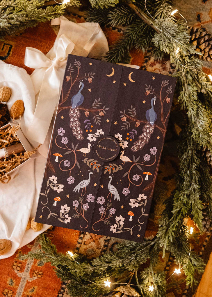 The "Advent Calendar - The Twelve Enchanted Days" by Mimi & August arrives in a decorative dark-toned gift box featuring elaborate peacock and floral patterns. It is accentuated with festive Christmas decor such as pine branches, walnuts, and cinnamon sticks. A white ribbon beautifully ties everything together on an elegantly patterned carpet, making it an ideal hiding spot for scented candles or a delightful advent calendar surprise!