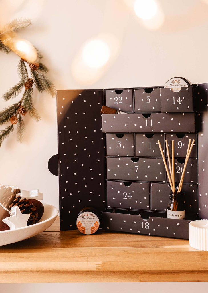 The Advent Calendar - The Twelve Enchanted Days by Mimi & August, featuring open drawers filled with scented candles and reed diffusers, sits on a wooden table adorned with pine cones and delightful Christmas decorations.