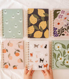 Six Mimi & August Agenda 2025 planners—featuring cups, lemons, flowers, peaches, cats, and blossoms—are arranged on a white surface. Two hands hold the cat design. Get yours FREE with the Agenda 2025 - Free Gift (100% off) offer!.