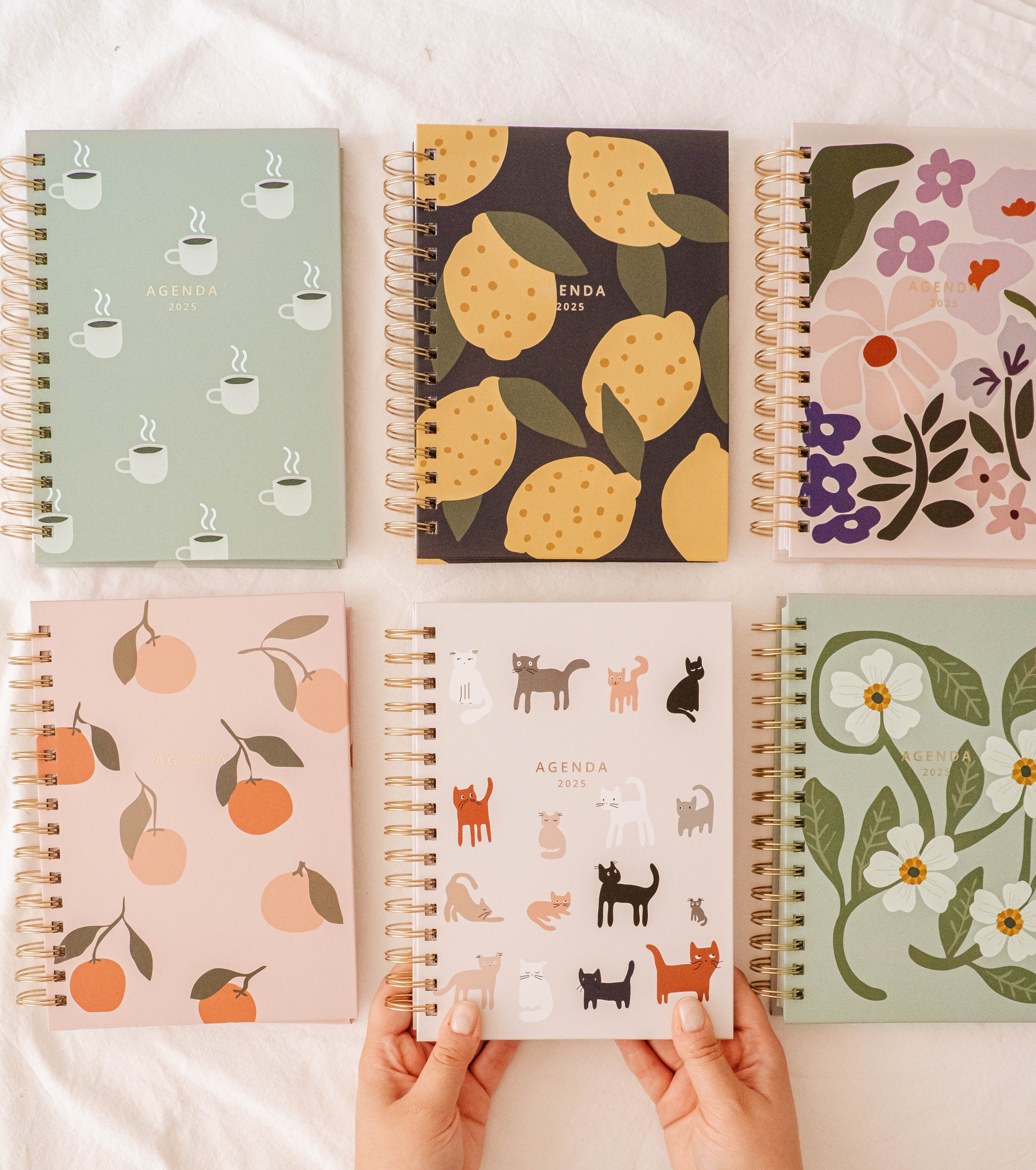 Six Mimi & August Agenda 2025 planners—featuring cups, lemons, flowers, peaches, cats, and blossoms—are arranged on a white surface. Two hands hold the cat design. Get yours FREE with the Agenda 2025 - Free Gift (100% off) offer!.