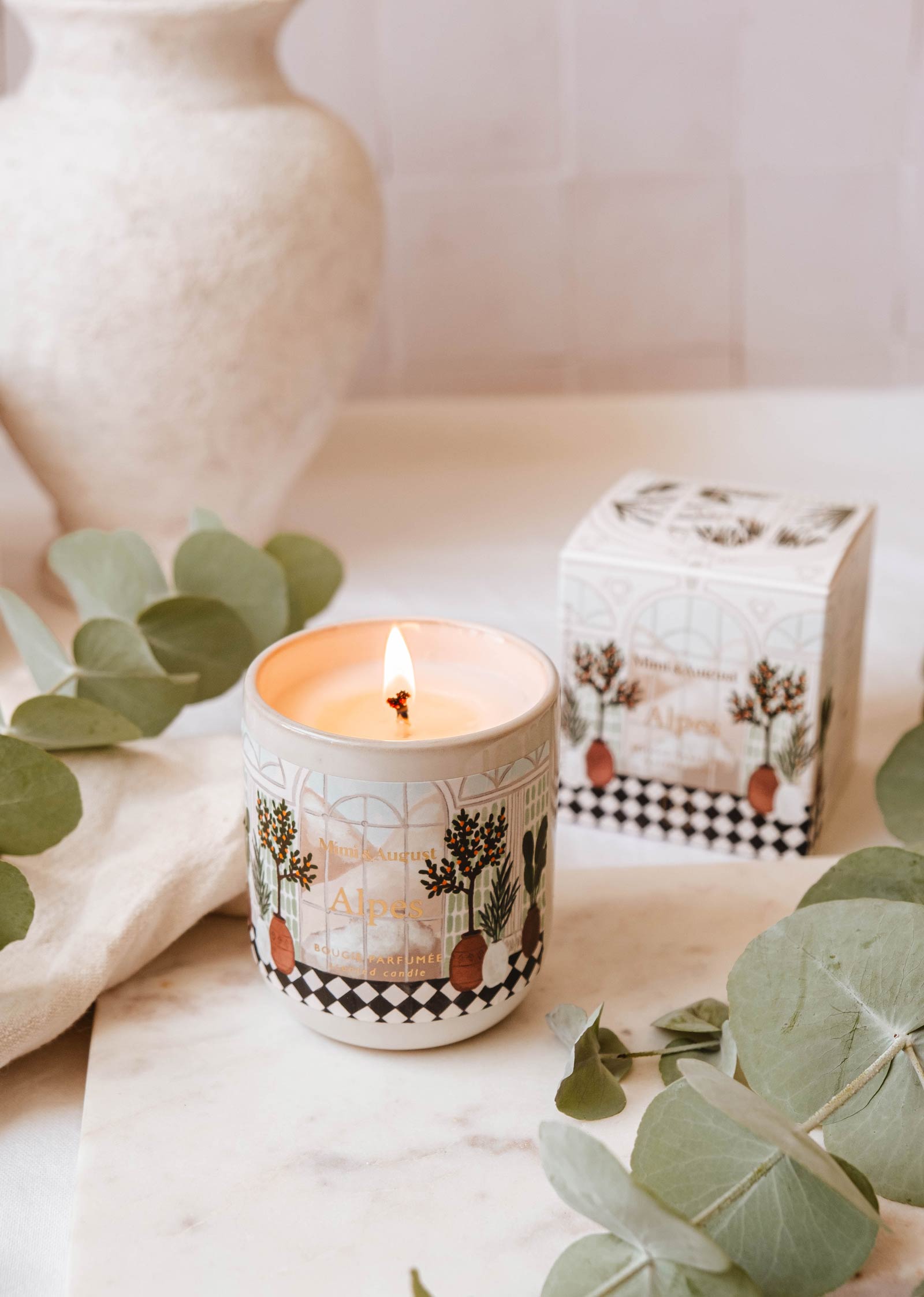 An Alpes - Reusable Candle by Mimi & August in a decorative ceramic container casts a warm glow on a marble surface near eucalyptus leaves, with its elegant reusable box positioned in the background.